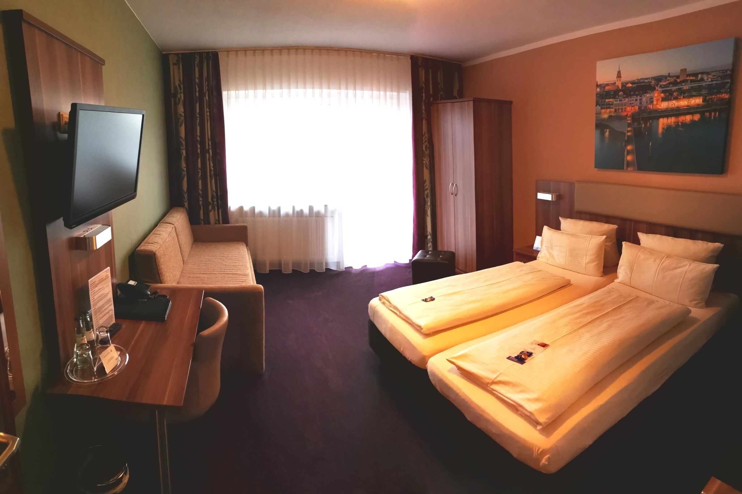 Une chambre d’hôtel avec un lit, un bureau, une télévision, une fenêtre avec des rideaux et un tableau au mur.
