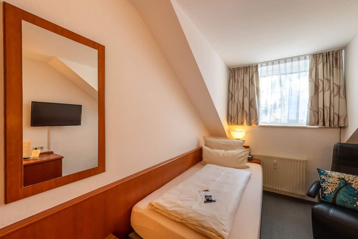 A hotel room with a bed, a chair, a TV mounted on the wall, a window with curtains, and a bedside lamp.
