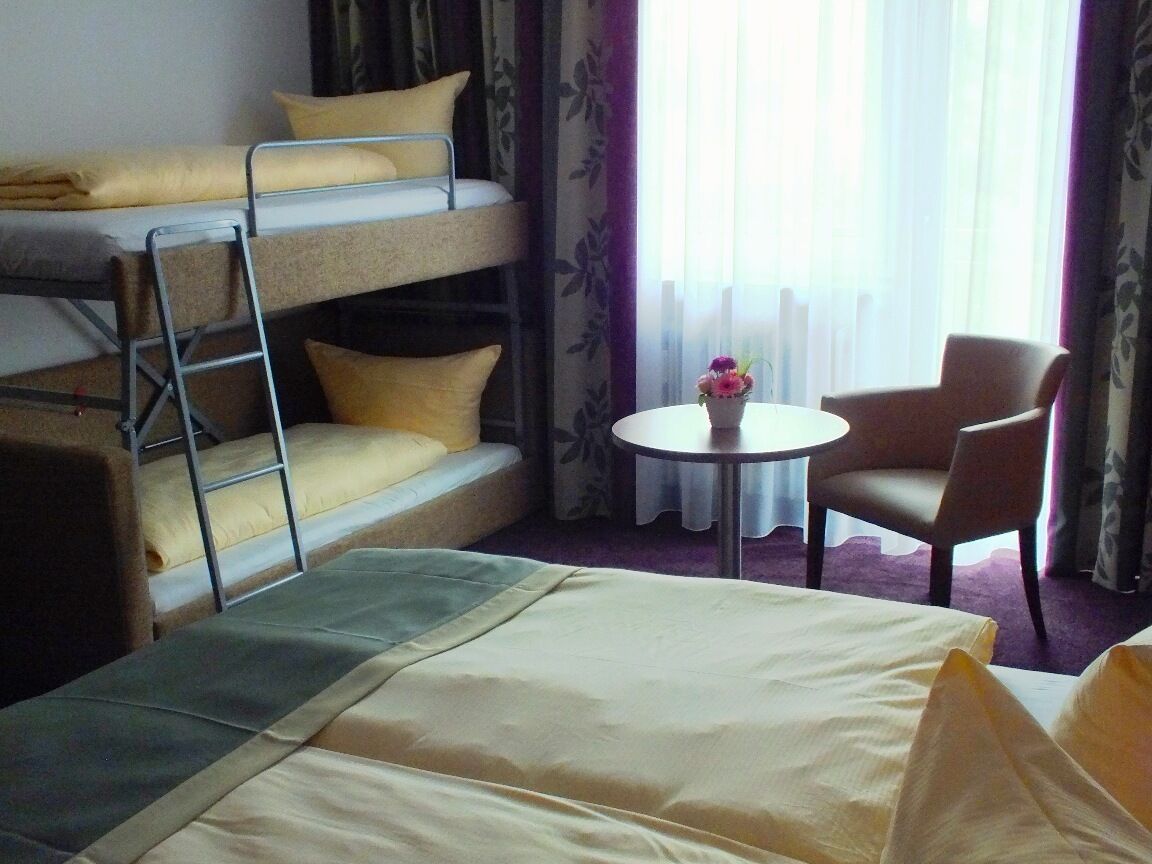 A hotel room with a bunk bed, a single bed, a round table with a vase, and a chair.