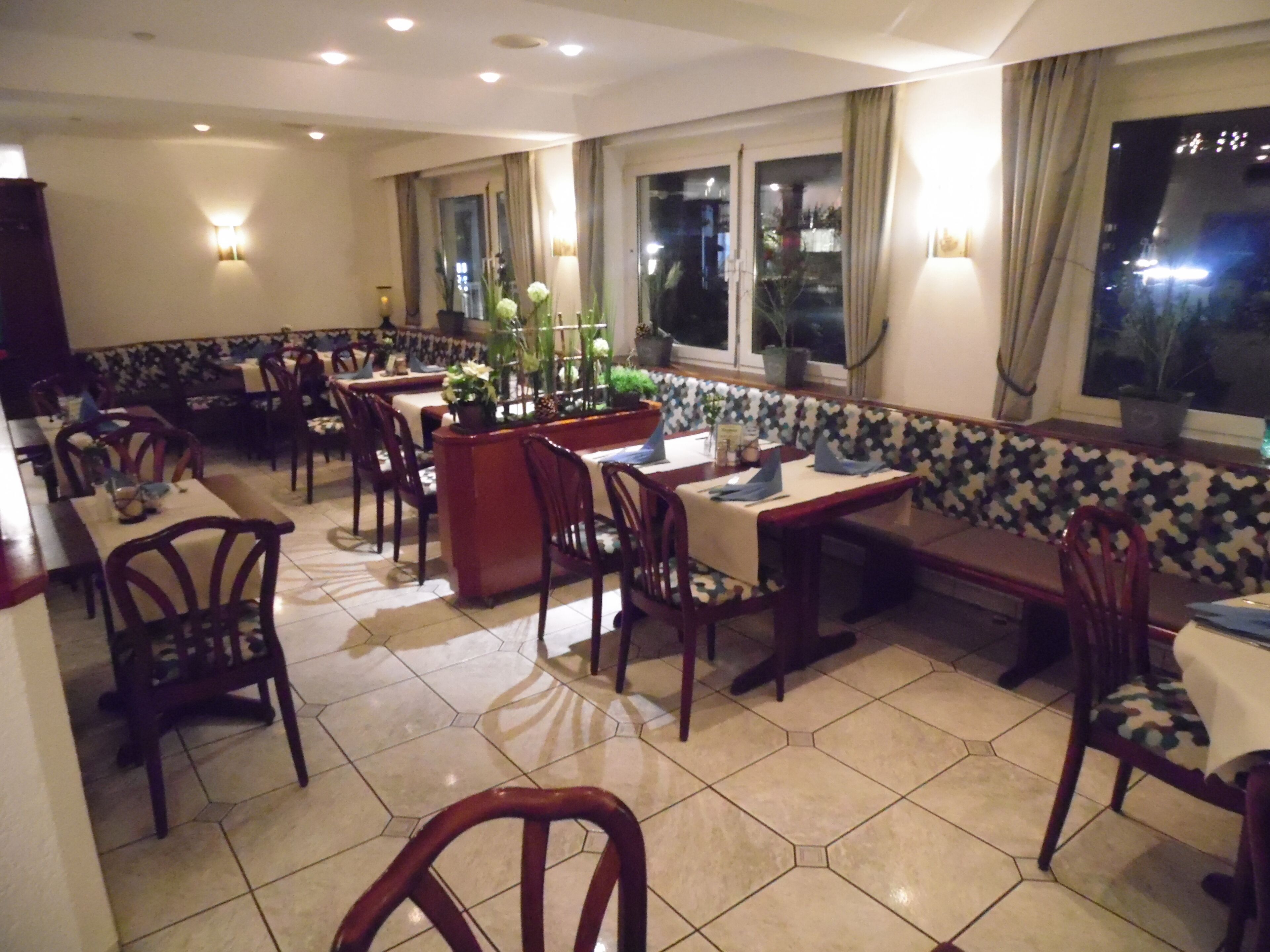 A restaurant with multiple tables and chairs, a central bar area, and large windows with curtains.