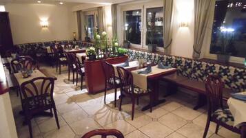 A restaurant with multiple tables and chairs, a central bar area, and large windows with curtains.