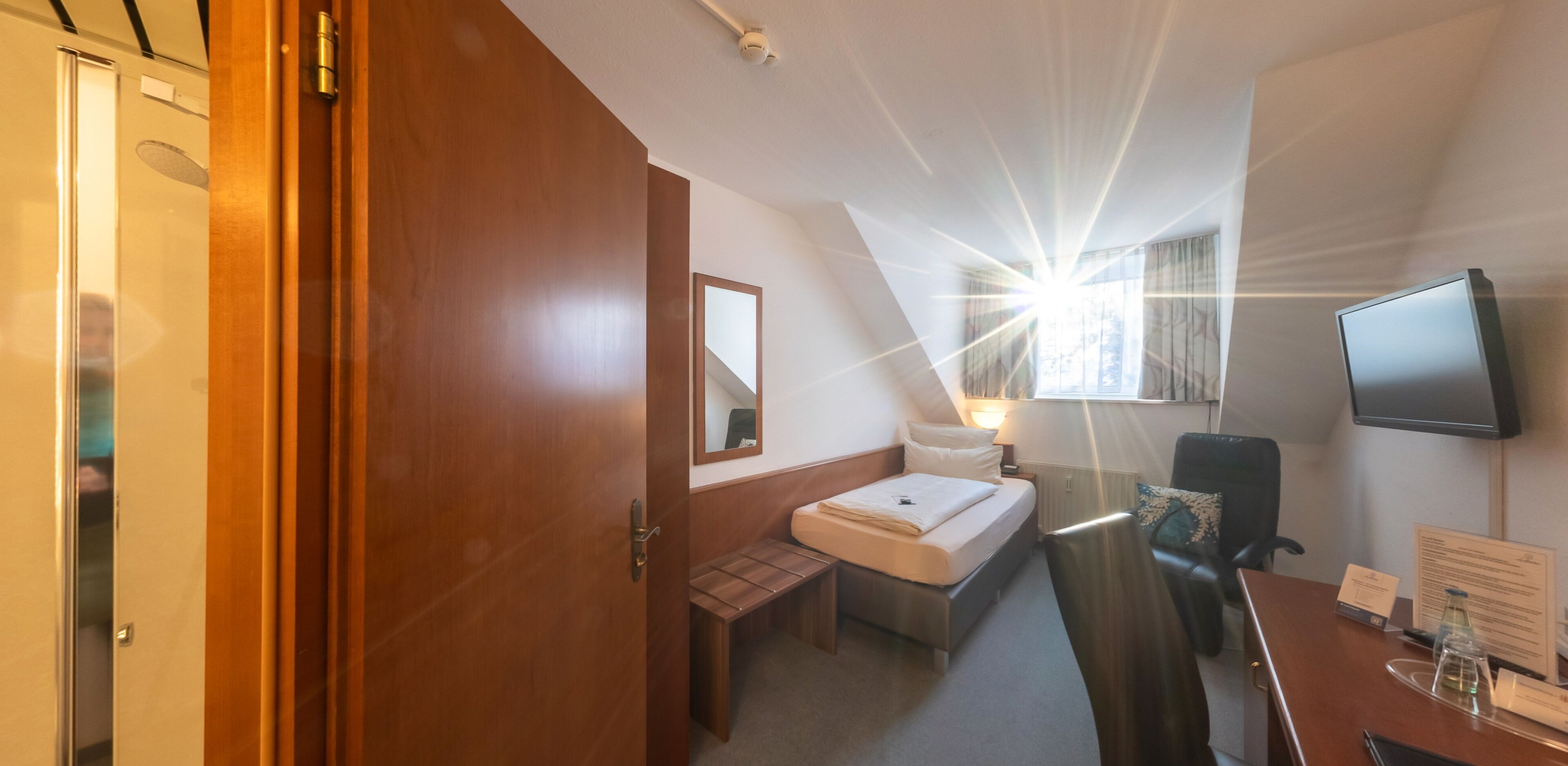 Habitación de hotel con cama, escritorio, silla, televisor y ventana con luz natural.