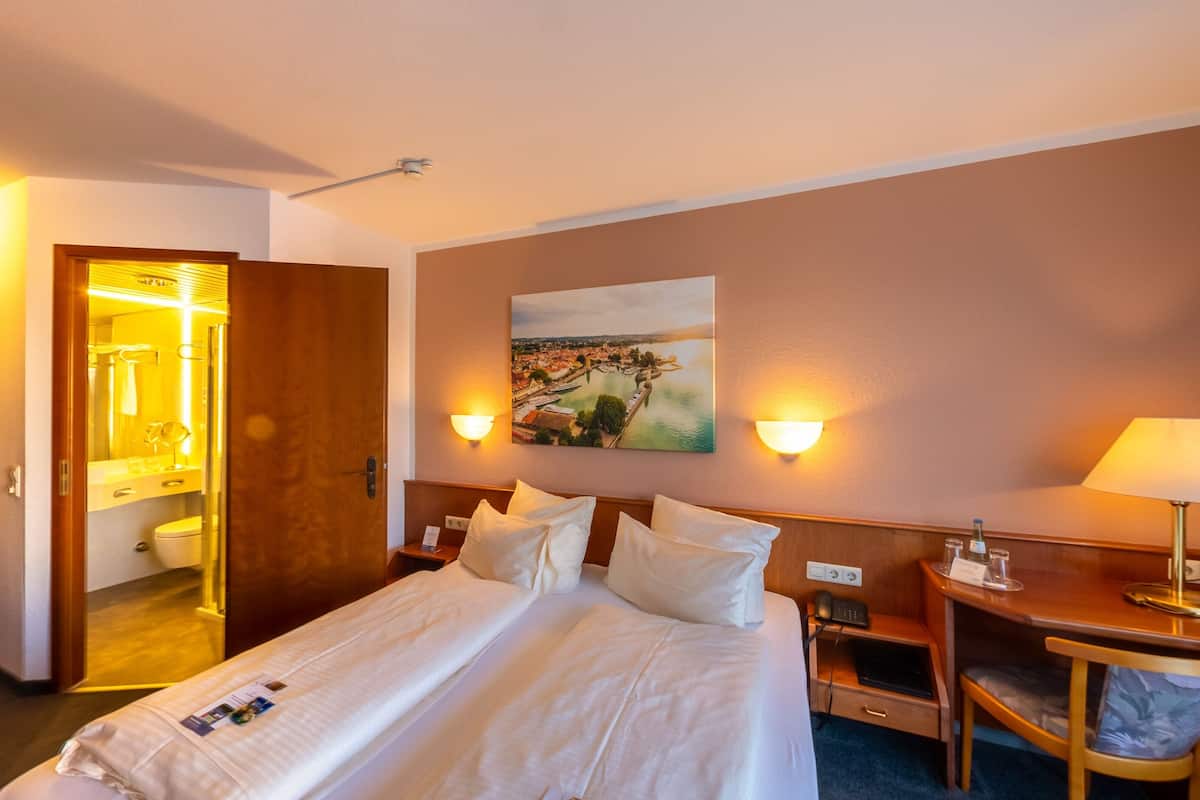 A hotel room with a bed, bedside tables, a desk, a chair, a painting on the wall, and a bathroom visible through an open door.