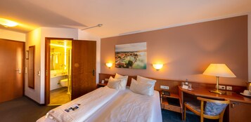 Quarto de hotel com cama, mesas de cabeceira, escrivaninha, cadeira, um quadro na parede e um banheiro visível por uma porta aberta.
