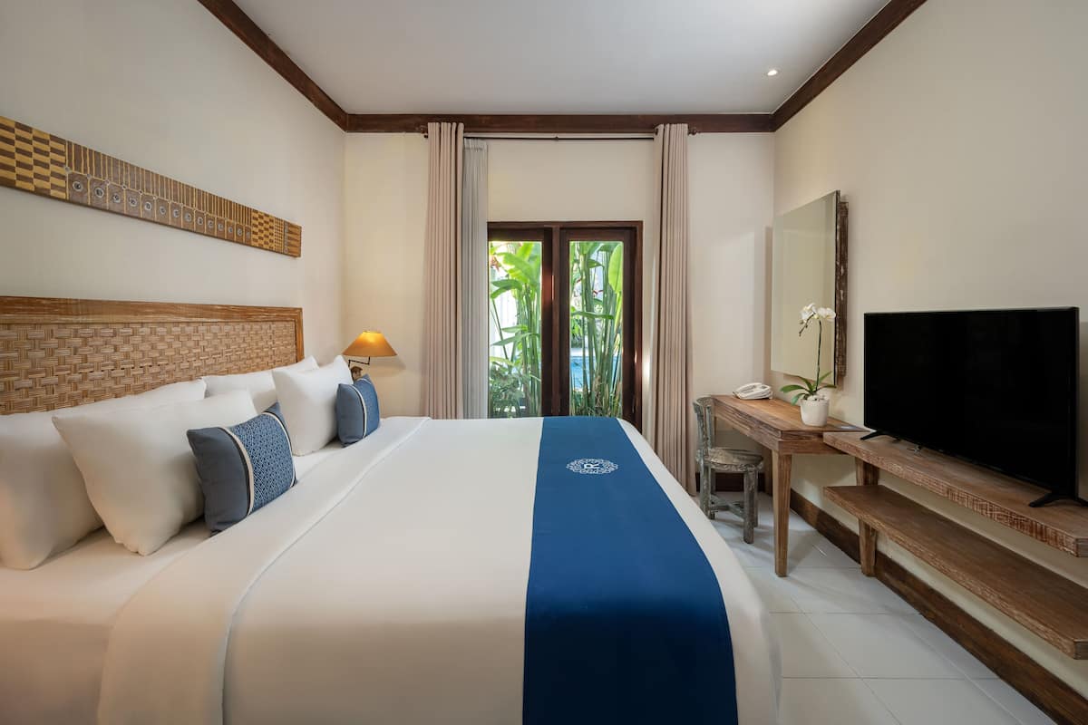 A hotel room with a large bed, a television, a desk with a chair, and a view of greenery through the window.