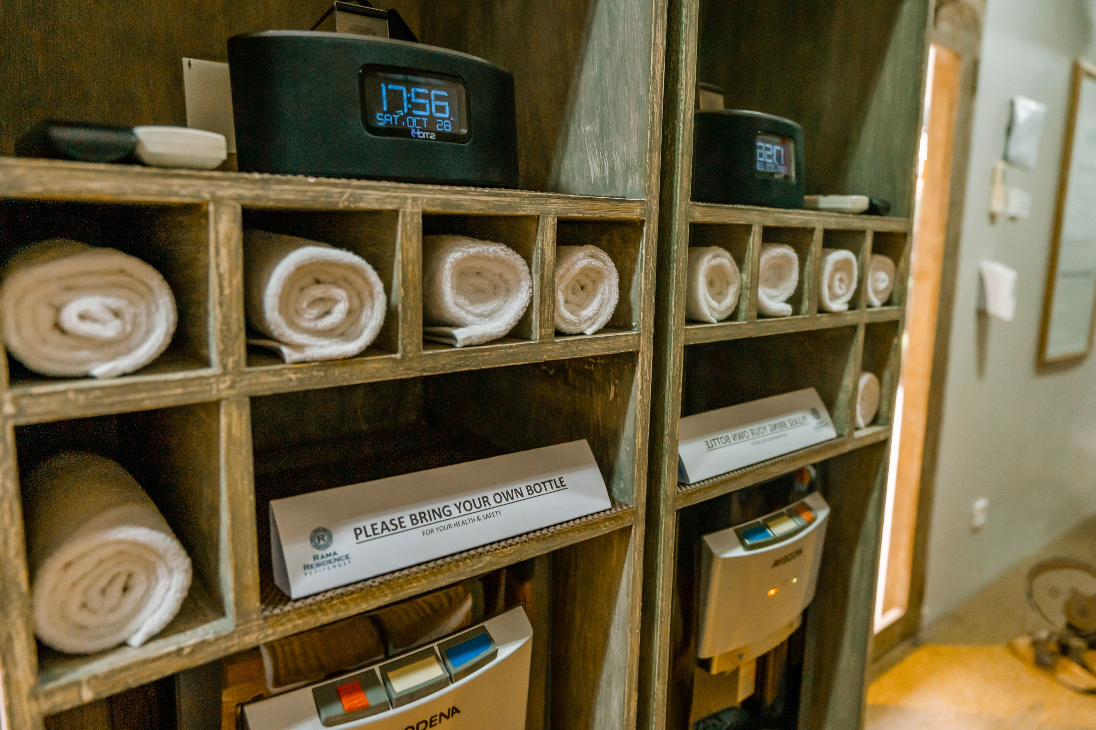 A hotel room with a wooden shelf displaying rolled towels and a sign instructing guests to bring their own bottle for health and safety.