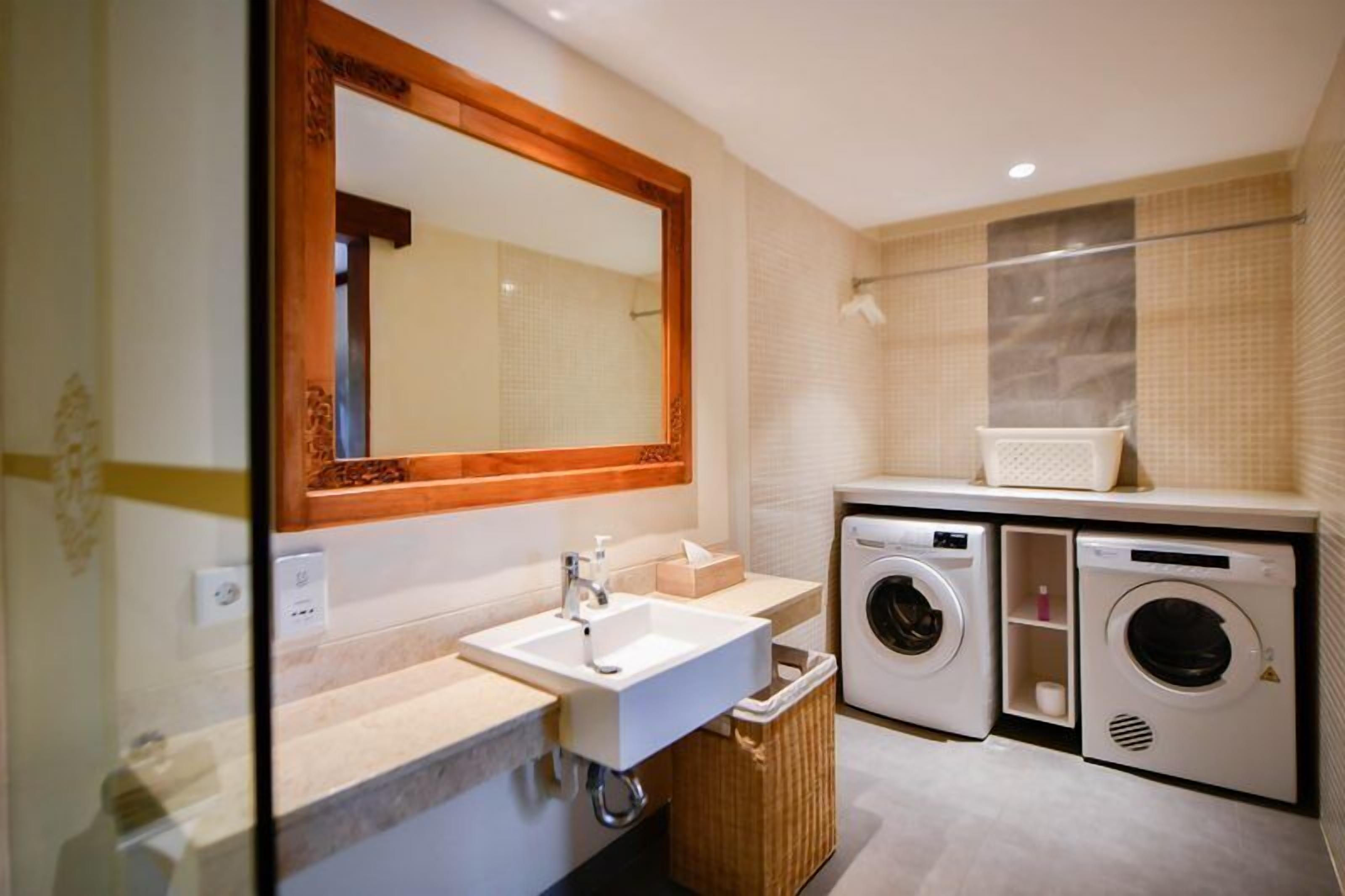 A compact bathroom with a sink, mirror, two washing machines, and a trash bin.