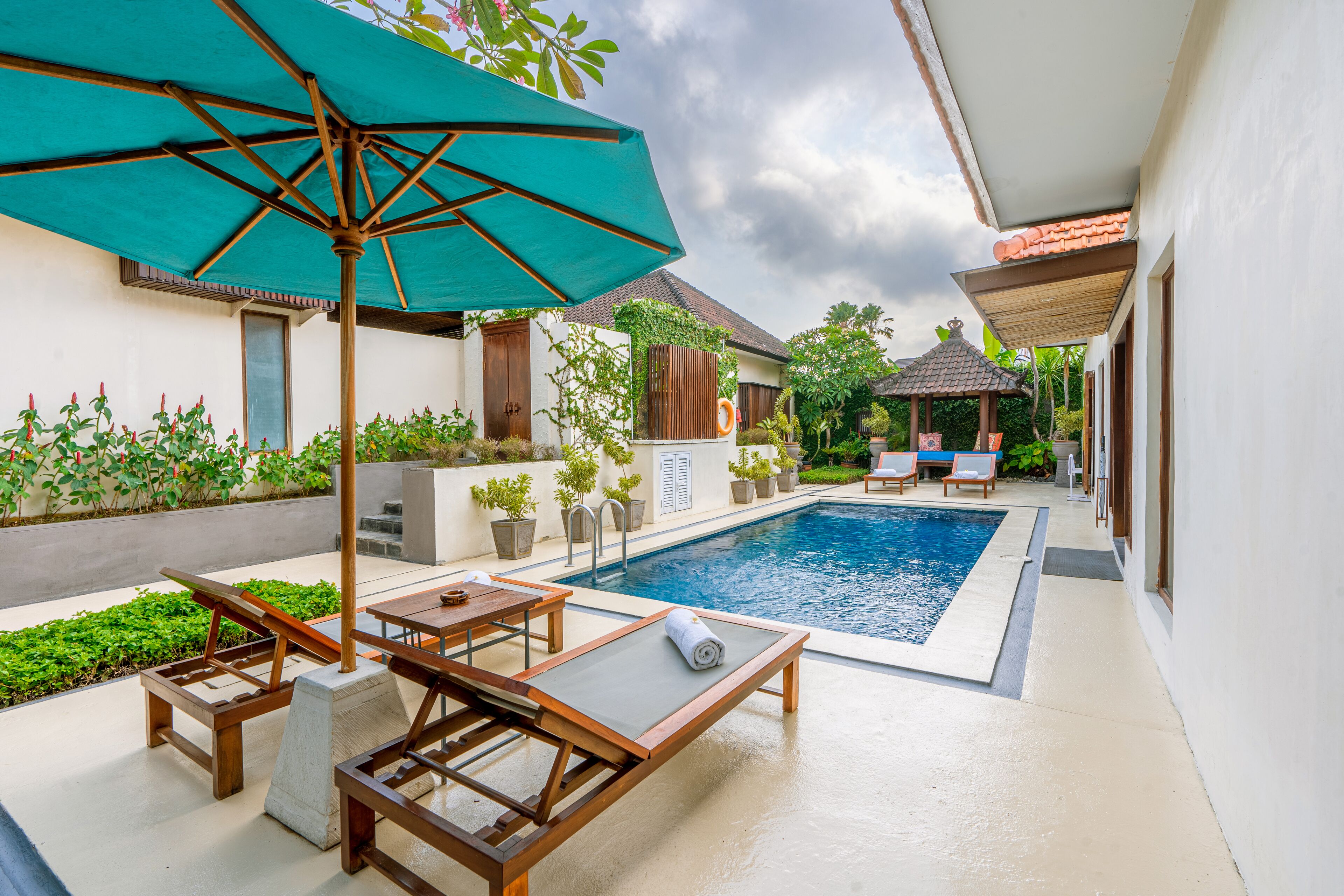 A swimming pool area with lounge chairs and a table, surrounded by potted plants and a pergola with a green umbrella.