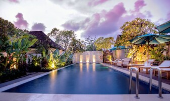 A swimming pool with lounge chairs and umbrellas, surrounded by tropical plants and a stone wall with water features.