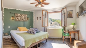 Un dormitorio con cama, una silla verde, una mesa de madera, un ventilador de techo y una ventana con vistas a un edificio y árboles.