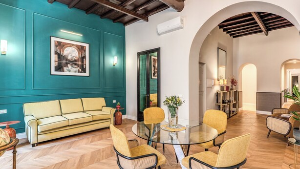 Una habitación con pared color turquesa, vigas de techo de madera, un sofá amarillo, una mesa de vidrio con sillas y un espejo.