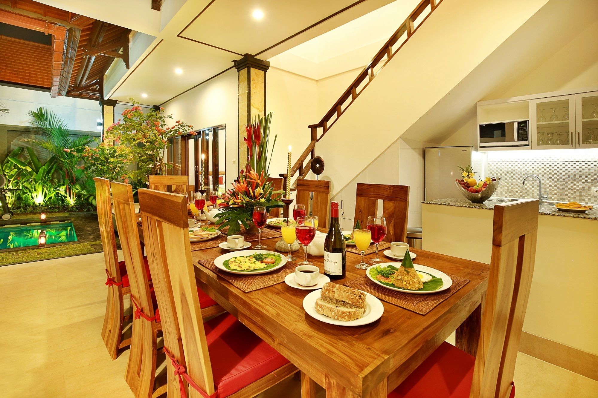 A dining area with a wooden table set for two, surrounded by red cushioned chairs. A small garden with a water feature is visible in the background.