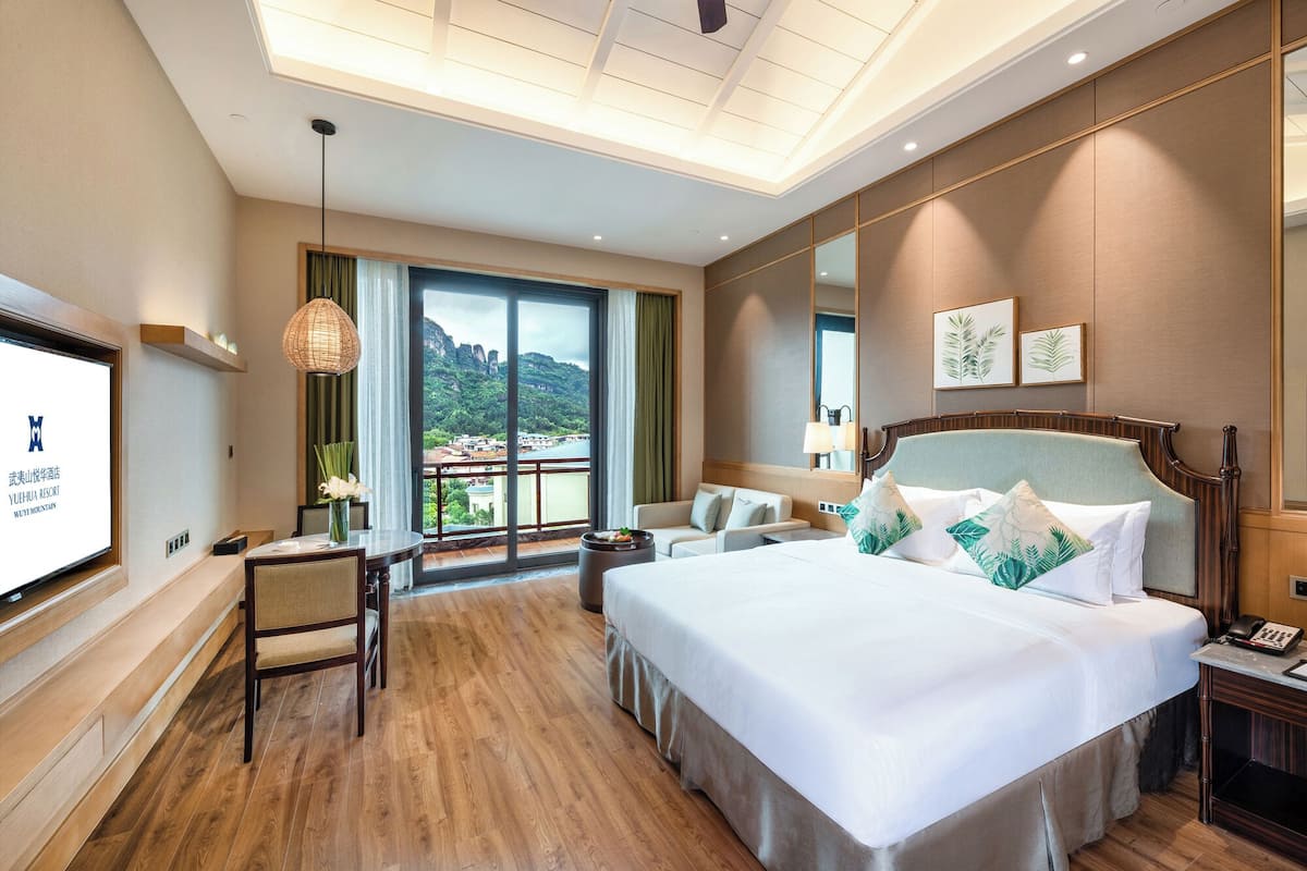 A hotel room with a large bed, a desk, a chair, a TV, and a balcony with a view of mountains.