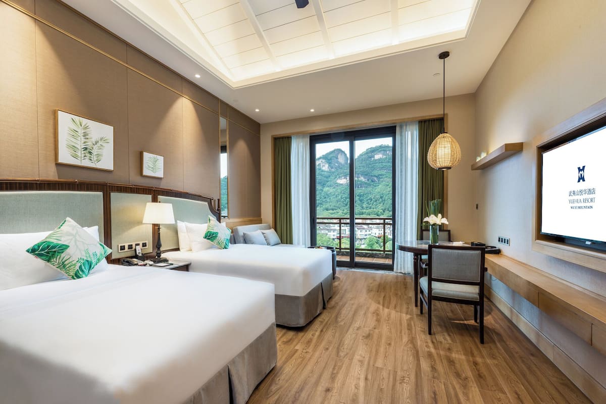 A hotel room with two beds, a desk, a chair, a large window with a view of mountains, and a ceiling light.
