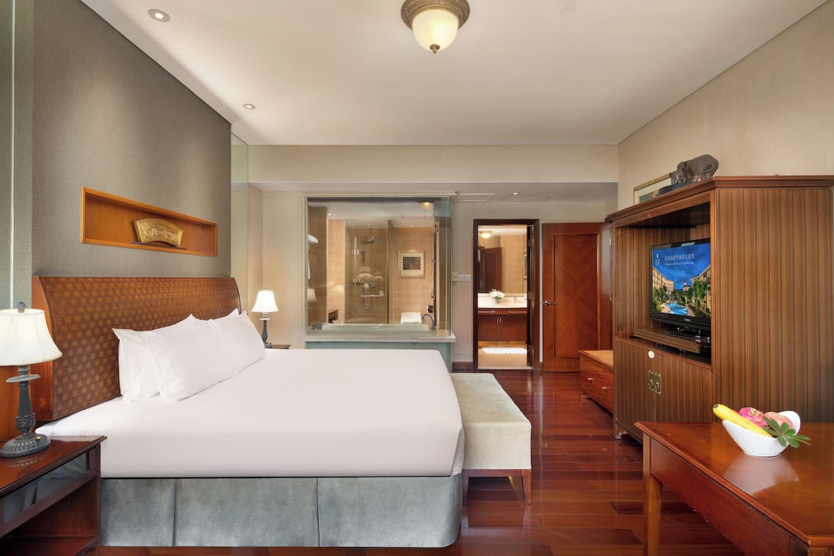 A hotel room with a large bed, bedside tables, a TV, and a bathroom visible through a glass door.