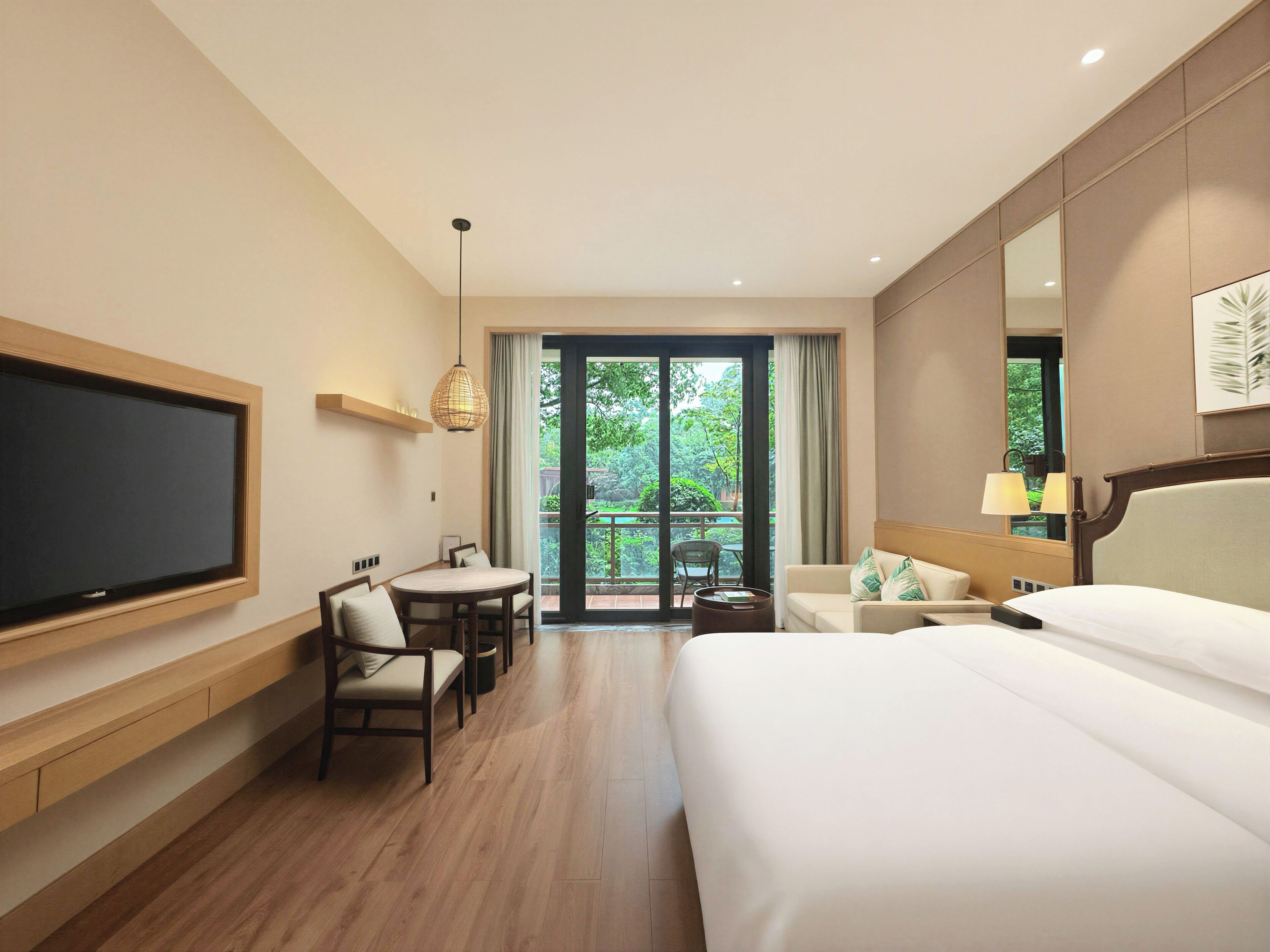 Une chambre d’hôtel comprenant un lit, une télévision, un bureau avec des chaises, un balcon donnant sur un espace verdoyant et un grand miroir.