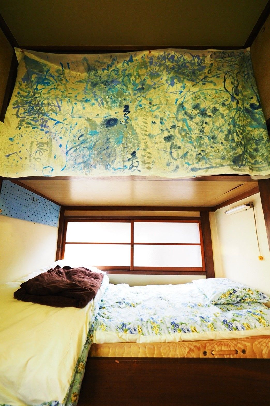 A dormitory room with bunk beds, a window with blinds, and a wall with a pegboard.