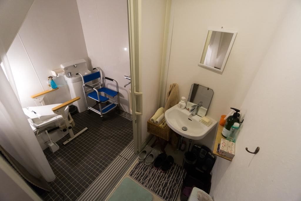 A small bathroom with a sink, mirror, and shower. There is a chair and a small table with a bottle of hand soap.
