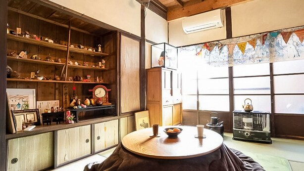 Una habitación japonesa tradicional con suelo de tatami, una mesa baja con un tazón y una taza, una radio y estantes con varios objetos.