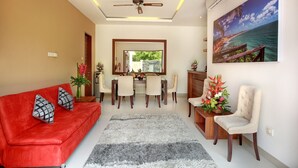 A modern living room with a red sofa, a dining area with a table and chairs, a ceiling fan, and a large window with a view of the ocean. - Lucia Villa Seminyak (Seminyak)