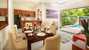 A modern dining area with a wooden table set for four, surrounded by beige chairs. A ceiling fan is visible above, and a large window offers a view of a swimming pool and greenery. - Lucia Villa Seminyak (Seminyak)