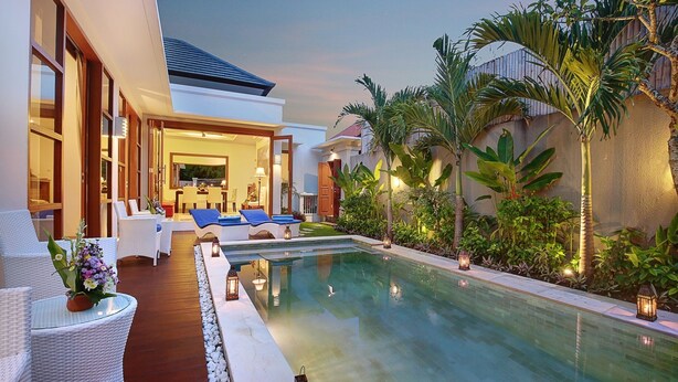 A poolside area with lounge chairs and a dining area, surrounded by tropical plants and a modern building. - Lucia Villa Seminyak (Seminyak)