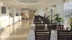 Un comedor amplio con mesas y sillas, una zona de cocina al fondo y ventanas grandes con plantas.