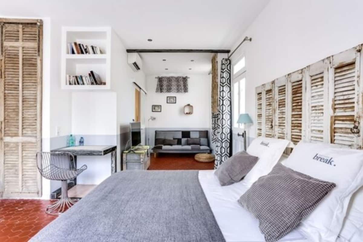 A bedroom with a bed, a desk, and a chair. There is a bookshelf and a window with shutters.