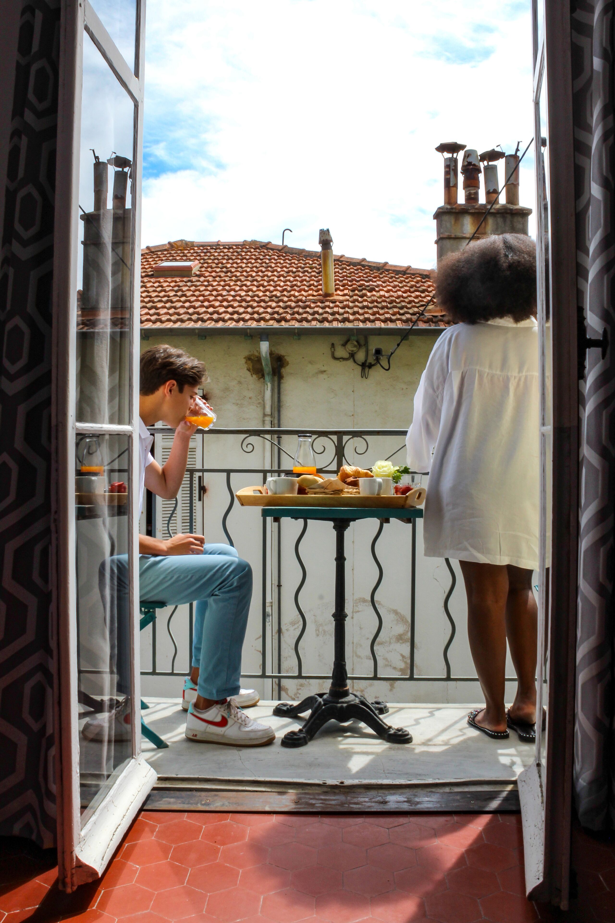 A balcony with a tiled roof, a table with food, and a person sitting and another standing.