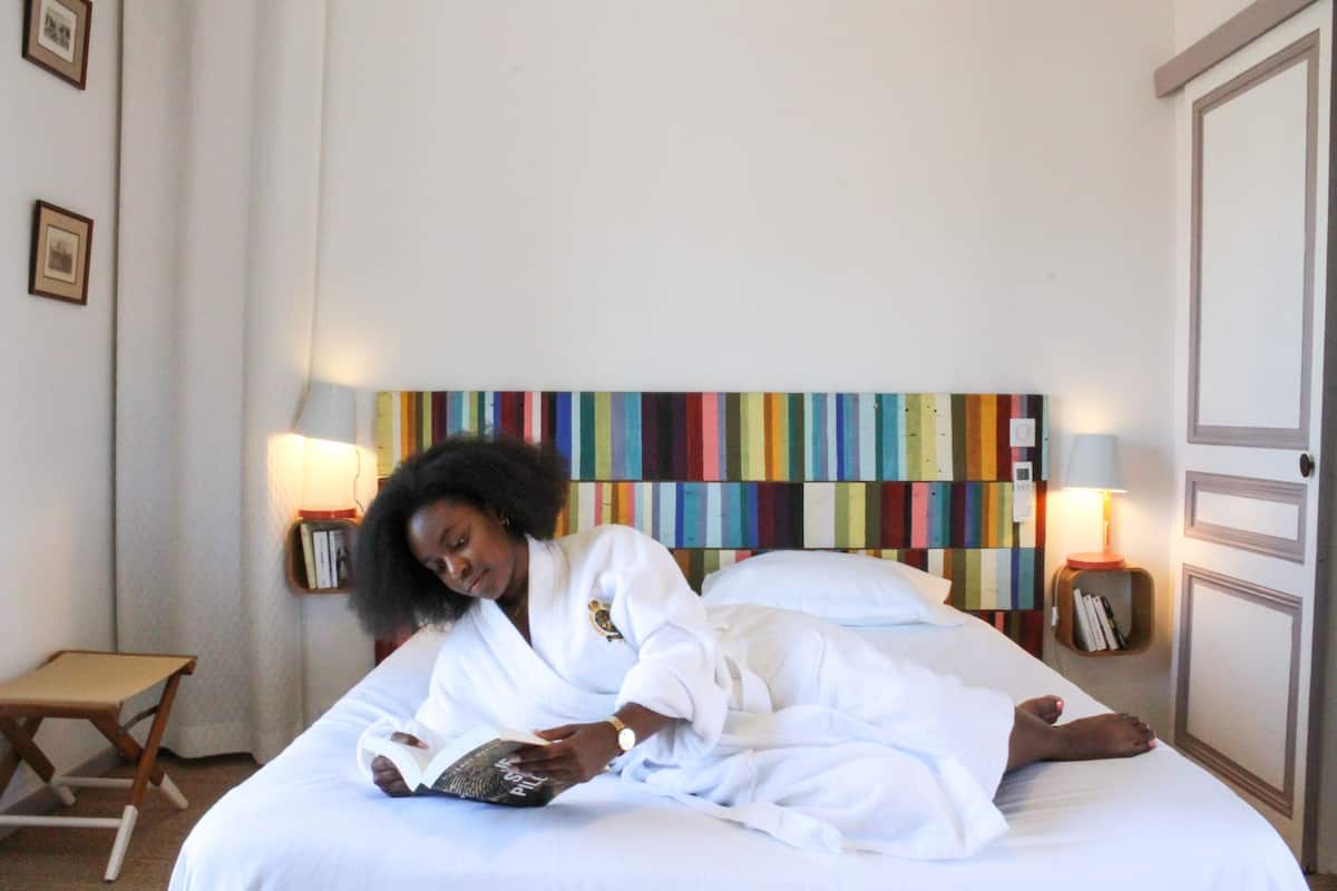 A person lying on a bed in a hotel room, reading a book. The room has a colorful striped headboard, bedside lamps, and a small wooden side table.