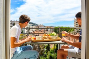 Two people sitting at a balcony table with a tray of food and drinks, overlooking a cityscape with buildings and greenery. - Villa du roc fleuri (Cannes)