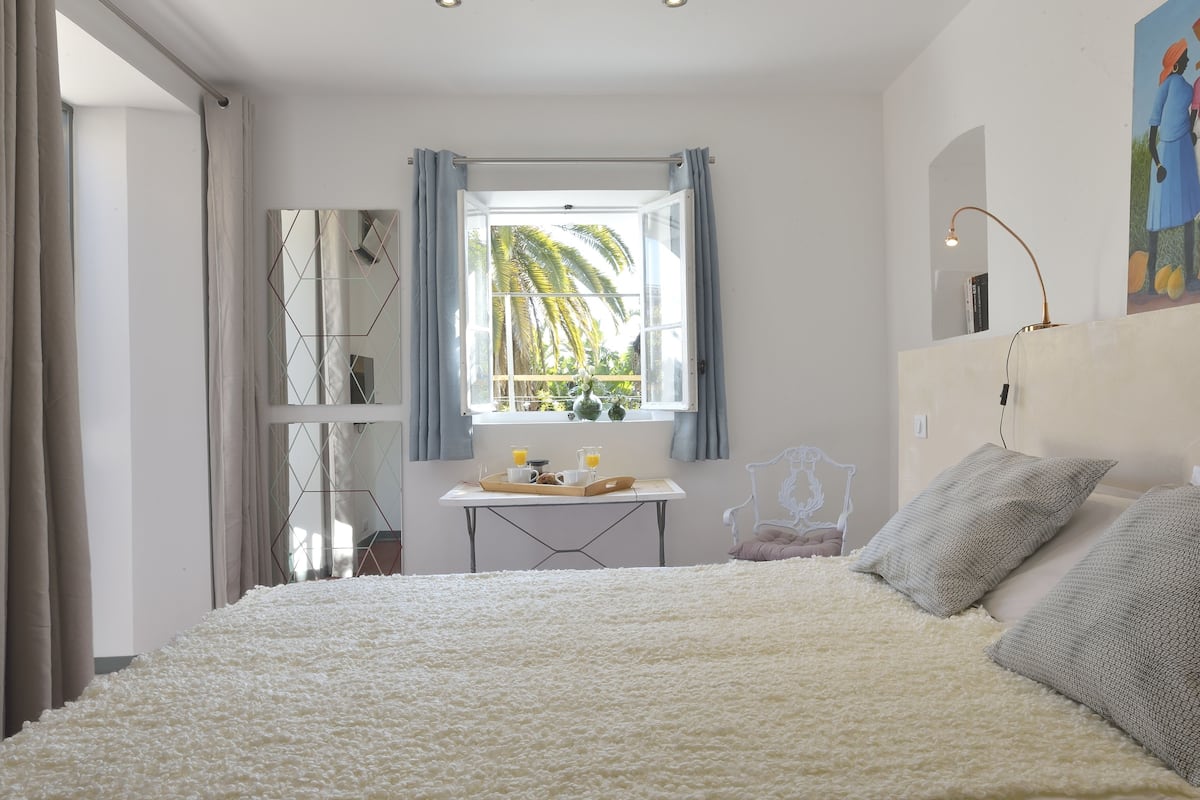 A bedroom with a large bed, a window with curtains, a small table with a tray of food, and a chair.