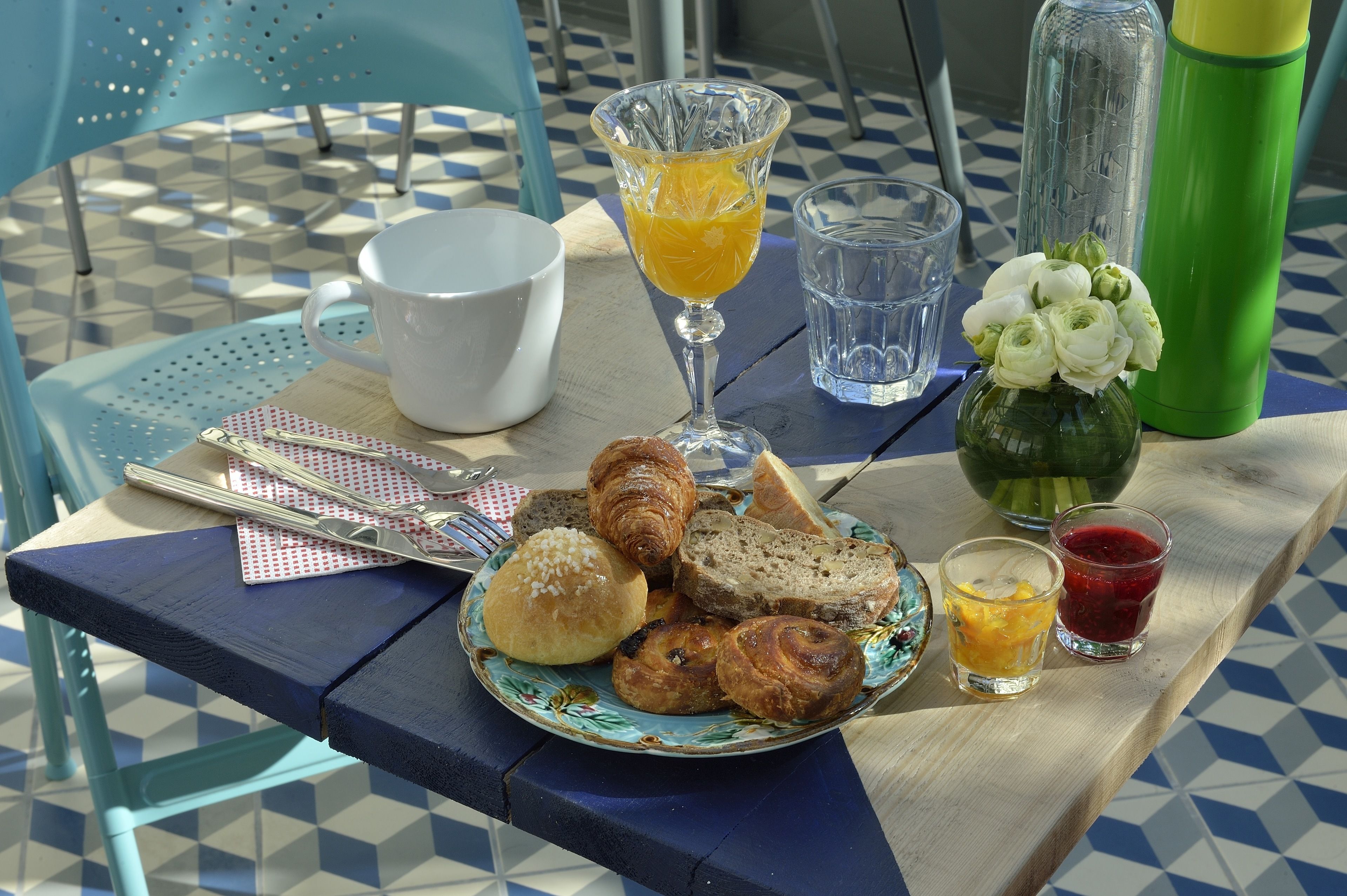 Una mesa de desayuno con cruasanes, pan, un plato de pasteles, un vaso de jugo de naranja, una taza de café, un vaso de agua y una botella de agua verde.