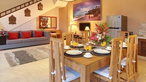 A dining area with a wooden table set for four, surrounded by wooden chairs. A sofa with red cushions is in the background, and a painting of a beach scene hangs on the wall. - Concetta Villa Seminyak Beachside (Seminyak)
