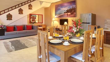 Un comedor con una mesa de madera dispuesta para cuatro personas, rodeada de sillas de madera. En el fondo, un sofá con cojines rojos y un cuadro de una escena de playa.