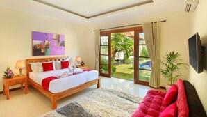 A bedroom with a large bed, a wooden headboard, a bedside table with a lamp, a TV mounted on the wall, and a view of the garden through a glass door. - Nicola Villa Seminyak Beachside (Seminyak)