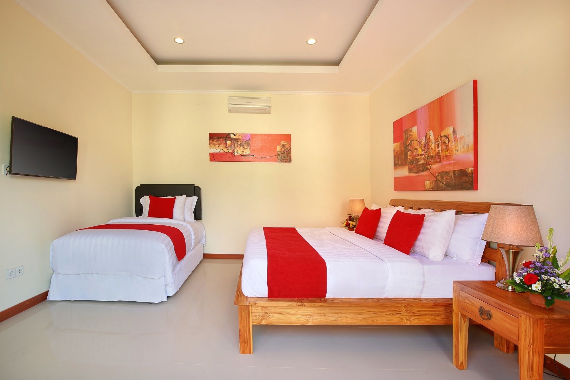 A hotel room with two beds, a TV, artwork, and a wooden nightstand with a lamp and flowers.