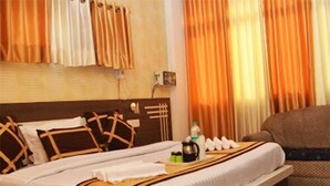 Quarto de hotel com uma cama grande, cabeceira de madeira, mesa de cabeceira com chaleira e pequena mesa com garrafa de água e uma chávena.
