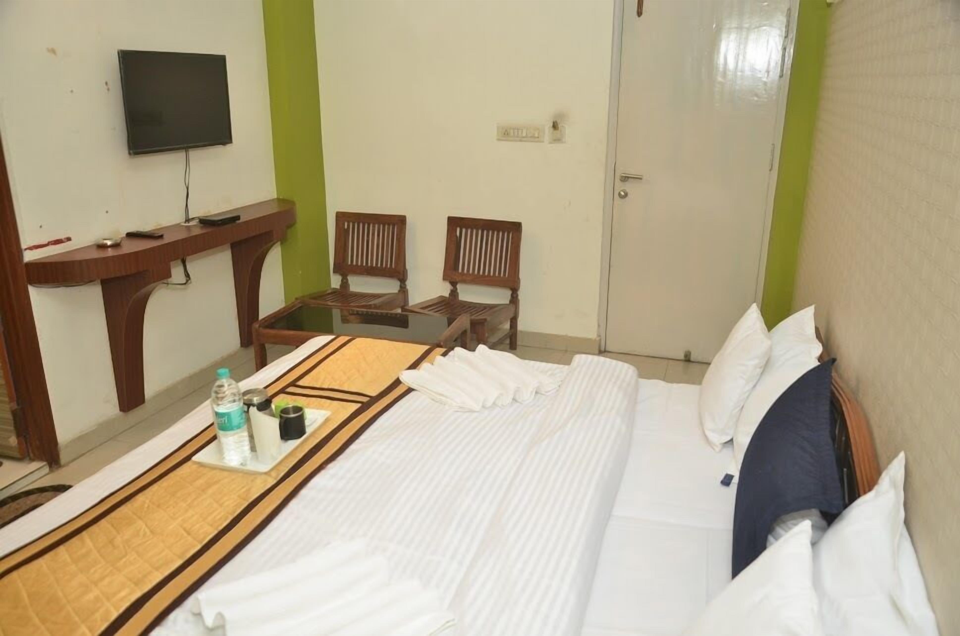 A hotel room with a bed, a TV mounted on the wall, two wooden chairs, a small table with a tray of toiletries, and a door.
