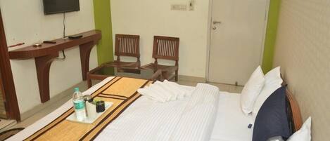 A hotel room with a bed, a TV mounted on the wall, two wooden chairs, a small table with a tray of toiletries, and a door.