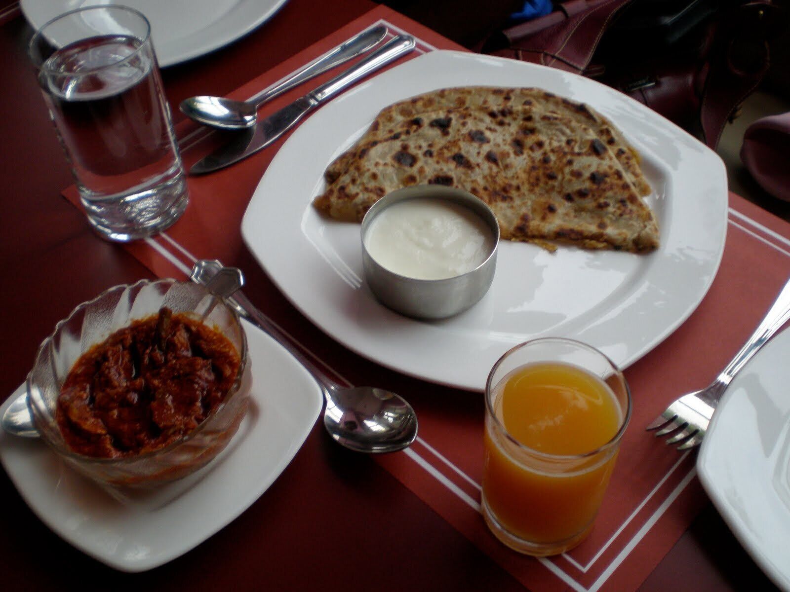 A meal with a flatbread, a bowl of yogurt, a glass of orange juice, and a bowl of chutney on a table.