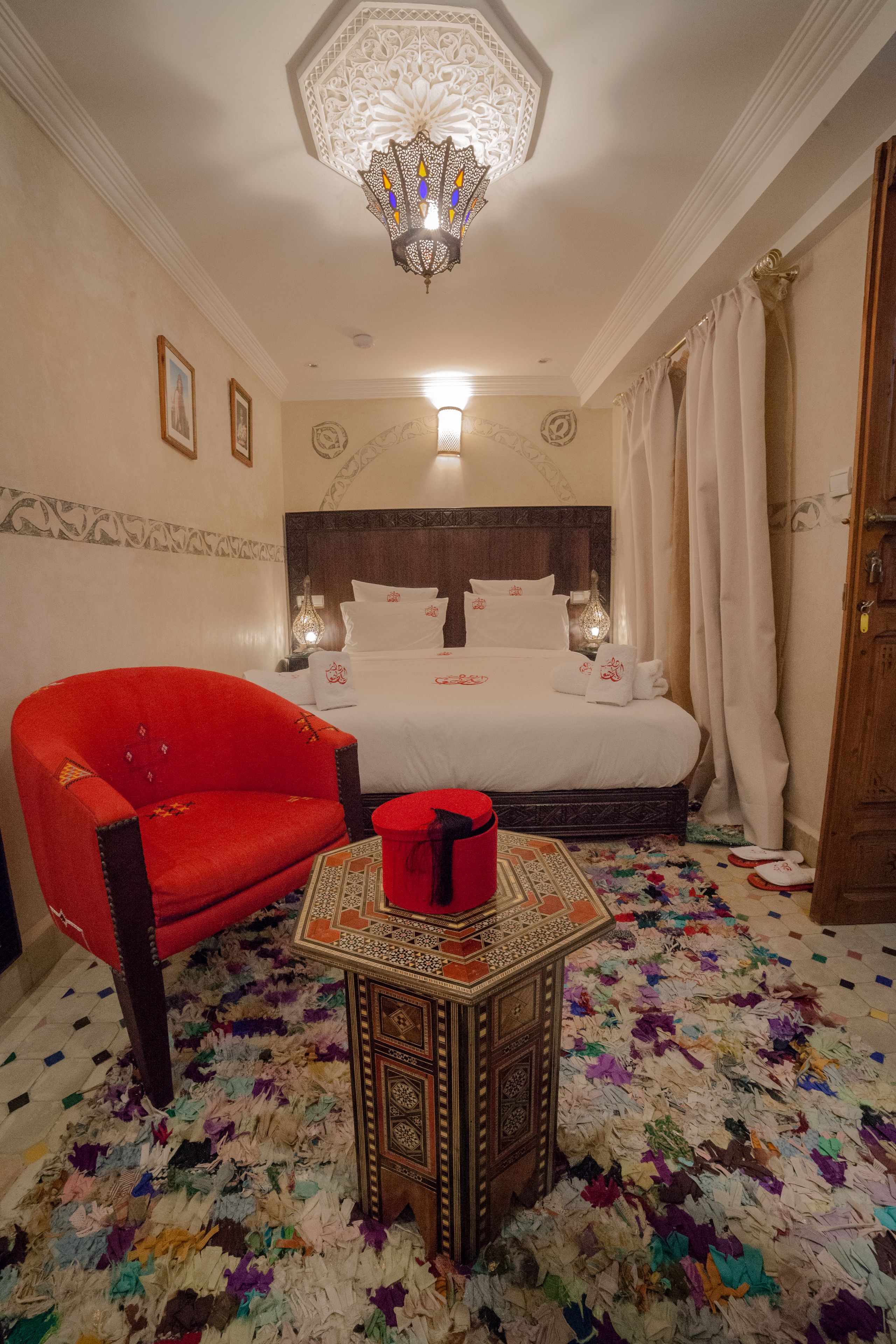 Habitación de hotel con una cama grande, un sillón rojo, una mesita con un objeto rojo y una alfombra estampada.