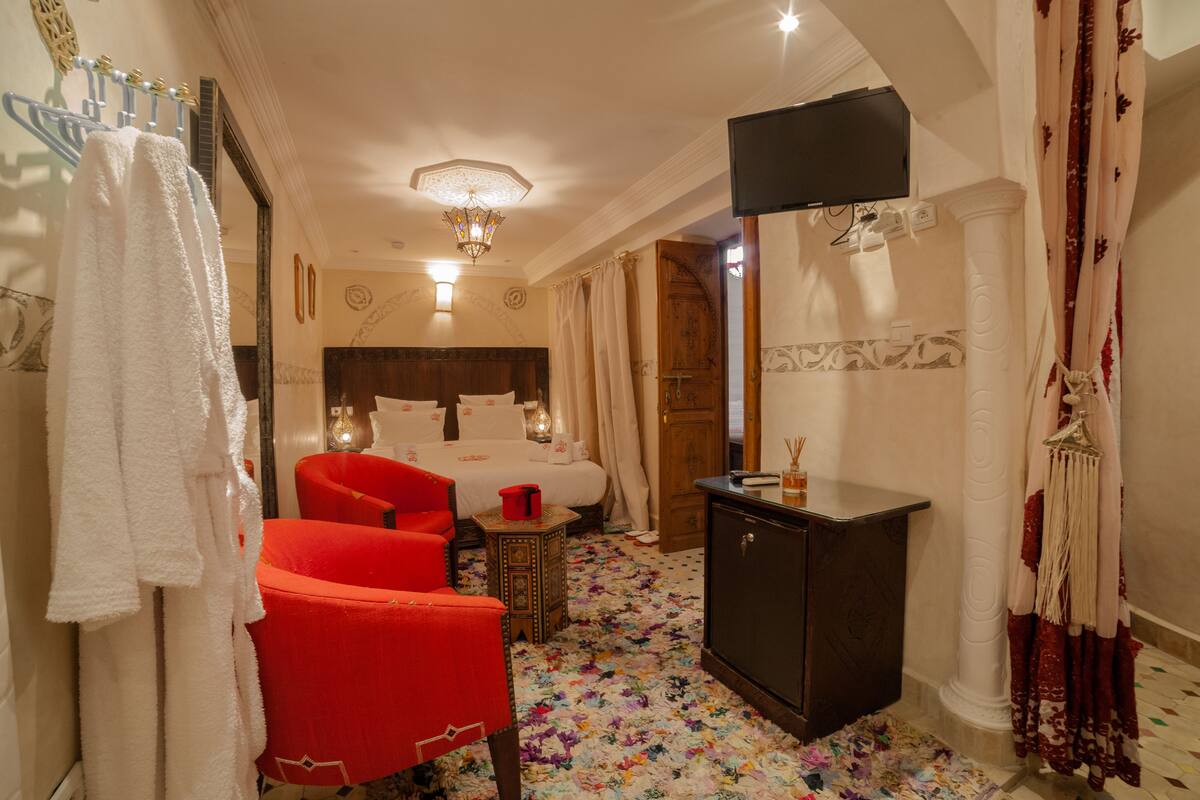 A hotel room with a bed, a red armchair, a small table, a wardrobe, and a television.