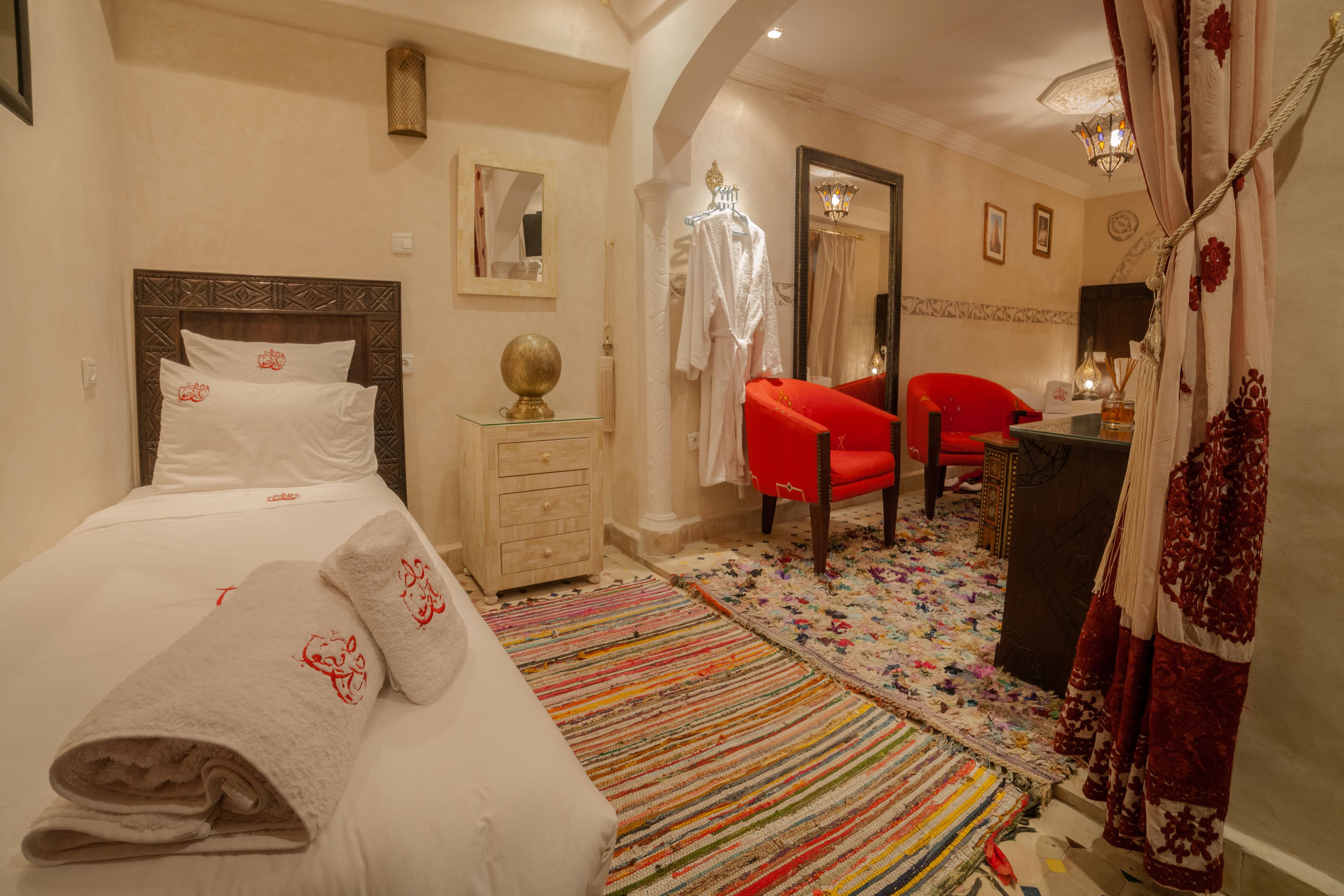 A hotel room with a bed, a dresser, a red chair, a mirror, and a colorful rug.