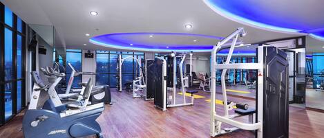 A modern gym with cardio machines, weight training equipment, and large windows offering city views.
