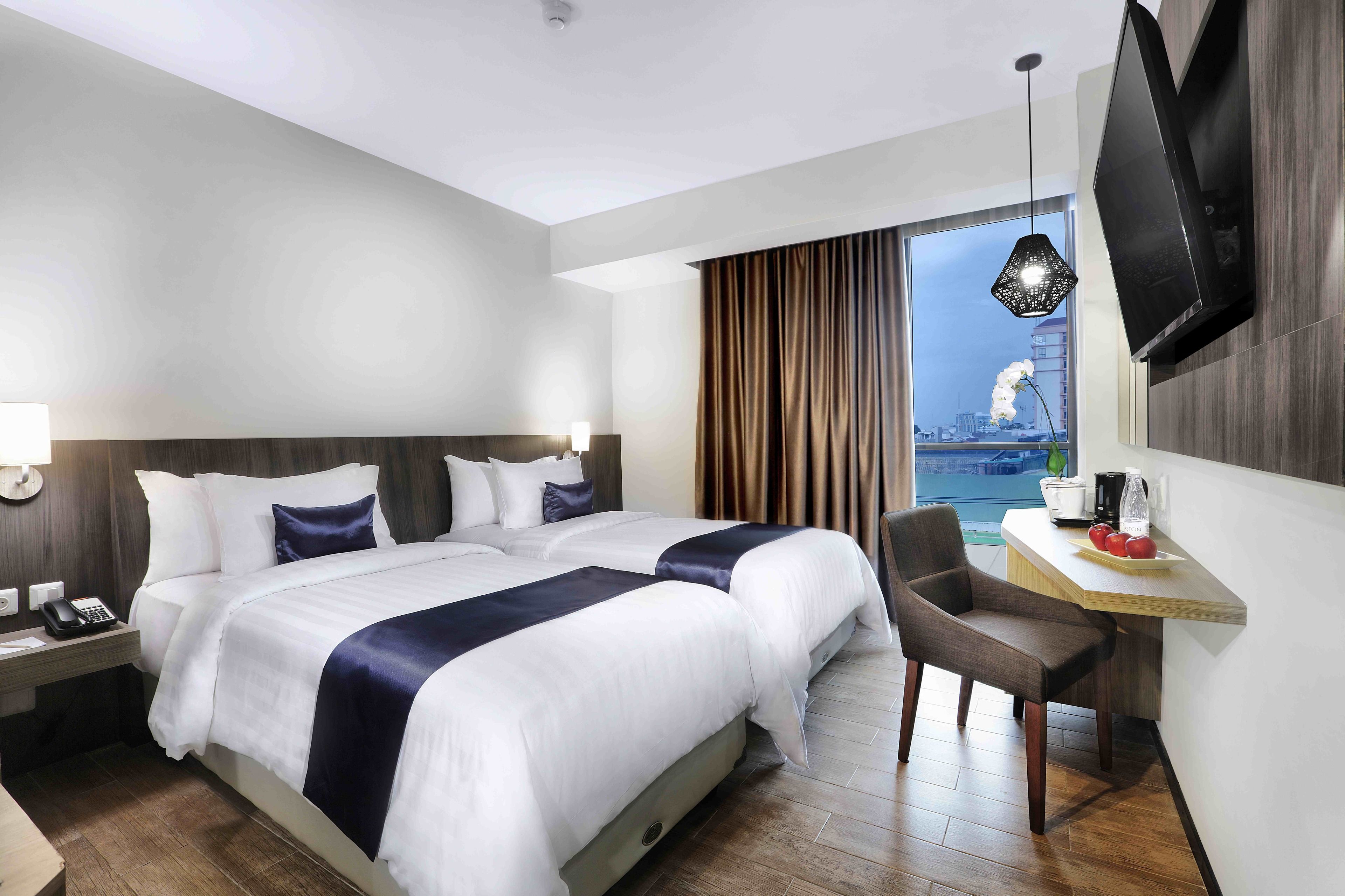 A hotel room with a large bed, a desk with a chair, a TV mounted on the wall, and a window with curtains.