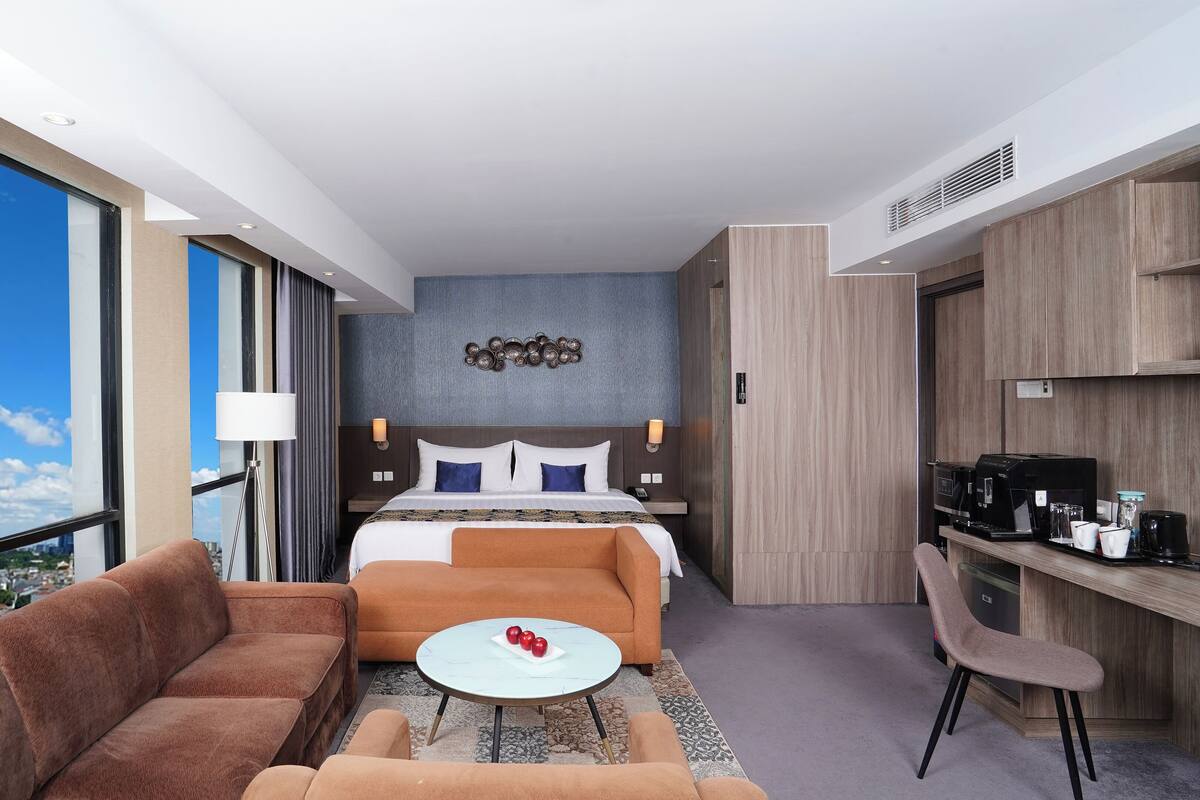 Ein Hotelzimmer mit einem großen Bett, einer Couch, einem kleinen runden Tisch, einem Schreibtisch mit einer Kaffeemaschine und Blick auf die Stadt durch große Fenster.
