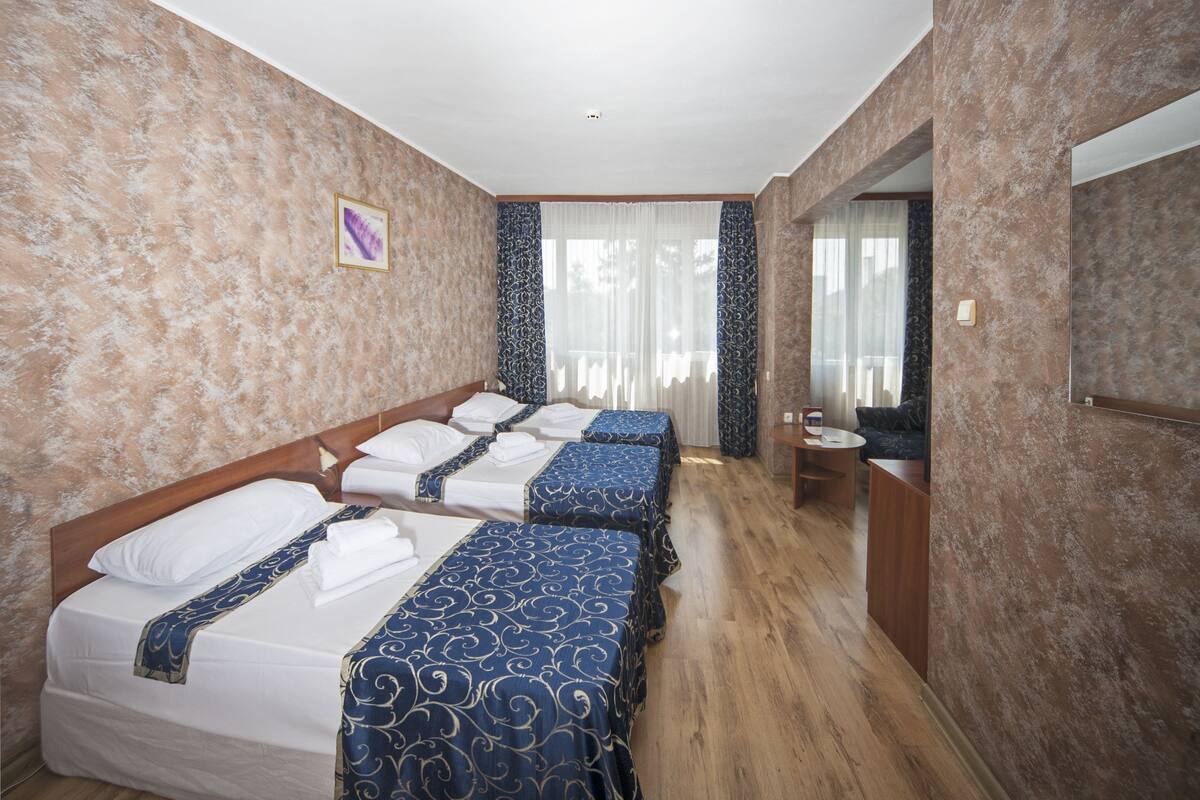 A hotel room with three beds, a wooden floor, a bathroom with a mirror, and a window with curtains.