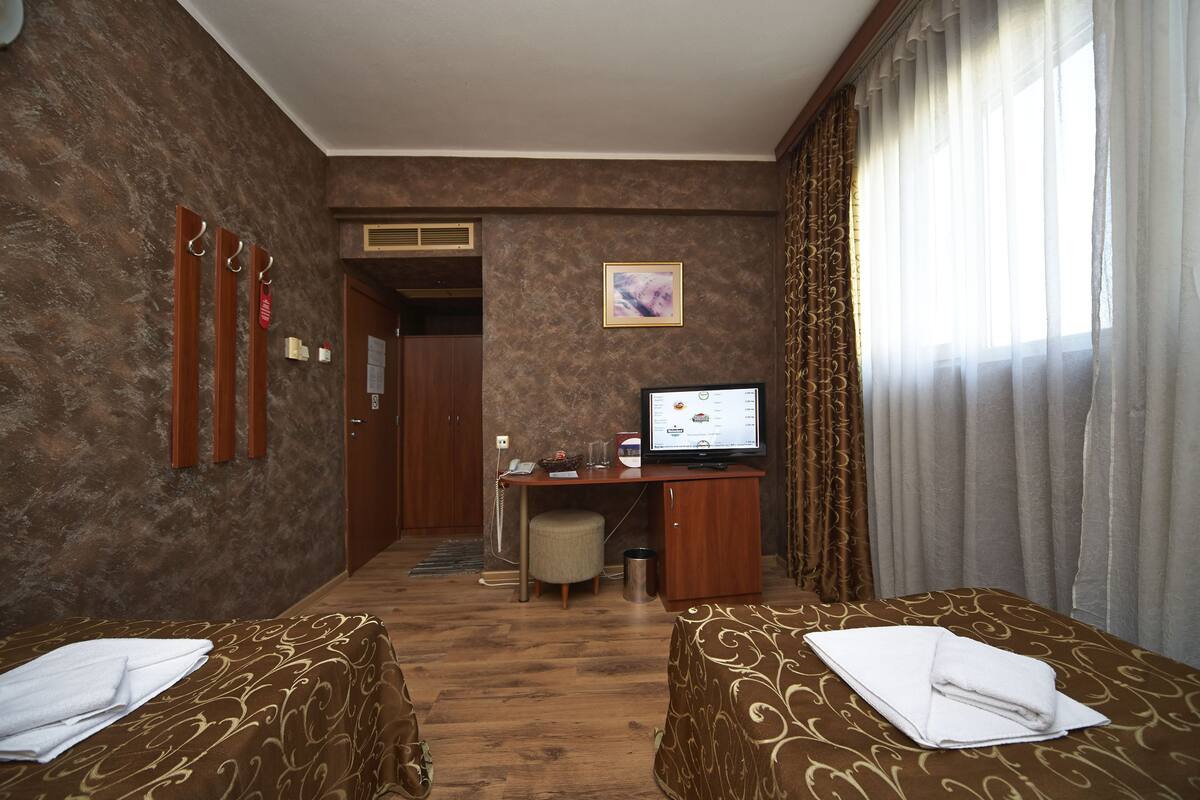 A hotel room with two beds, a desk with a computer, a small trash bin, and a window with curtains.