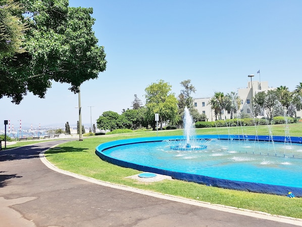 A circular fountain with water jets in the center, surrounded by a blue pool, set in a park with grass and trees. - Carmel Apartments (Haifa)